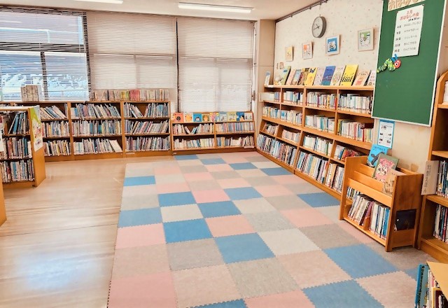 壁際にある木製の低い本棚に置かれた本たちと、カラフルなじゅうたんが敷かれたえほんコーナーの写真