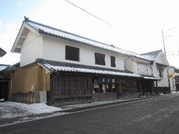 道路沿い、のれんがかかっている店舗の写真。入口は木造、二階は白のしっくい壁、瓦屋根の建物。蔵も隣接している。