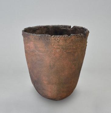 土器の写真。赤褐色に近い茶色、卵を半分に割ったような形の入れ物で、先端はとがっていない。模様などはないが、上部に線のような模様がある。