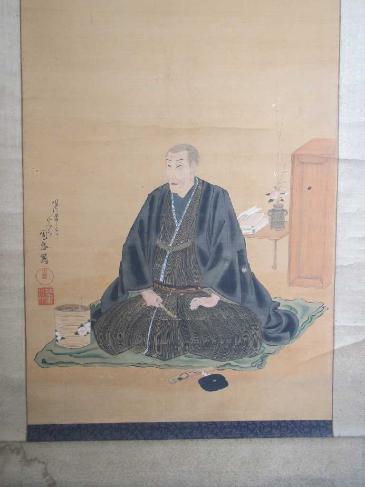 掛け軸に描かれた初老の男性。着物姿だが、髪の毛は五分刈りにされている。手前にキセル、脇に小さな火鉢がある。