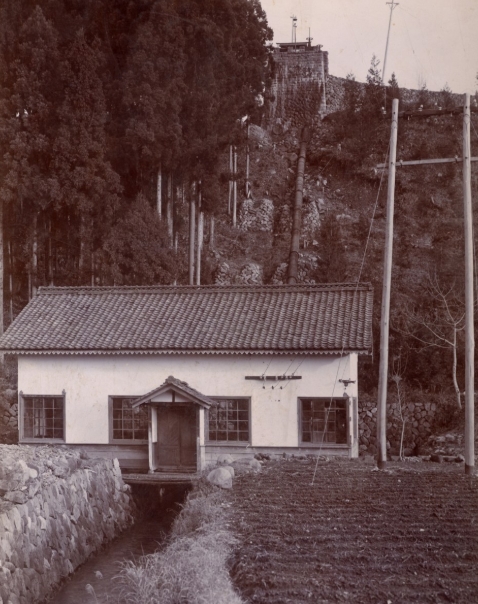 セピア色のモノクロ写真、明治37年に設立した当時の発電所写真。平屋で瓦屋根、建物の下を川が流れている。