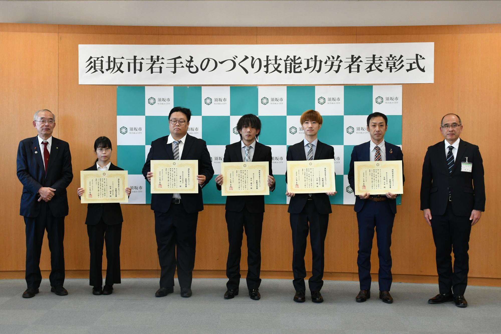 表彰状を胸の高さに持った5人の須坂市若手ものづくり技能功労者と須坂市長と産業振興部長が、横断幕の前に並んで記念撮影している様子