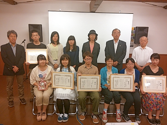 一列目に額に入った表彰状を持ち椅子に座っている人6名、2列目に7名の方が立っている総勢13名の集合写真