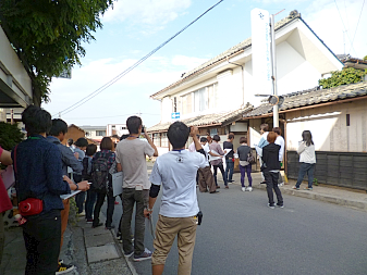 道路を挟んだ先に、白い縦看板が設置された白い壁の建物があり、それを大勢の信州大学工学部建築学科の生徒達がカメラで撮影などしている写真