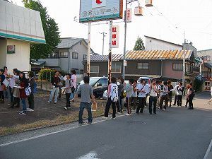 市街地の道路に面し「駐車場」と書かれた看板が設置されたスペースに大学生が集まっている写真