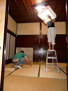 和室の照明器具を脚立に乗って取り外す大学生の写真
