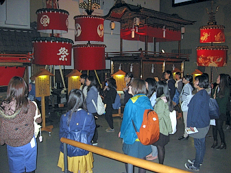 うす暗い中、白文字で祭りと描かれた赤い笠鉾や立て札、山車が展示され、学生の方々が見学している様子の写真