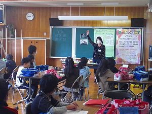 黒板の前で手を挙げて話をしている女性とグループごとで席に座った子どもたちが話を聴いている写真