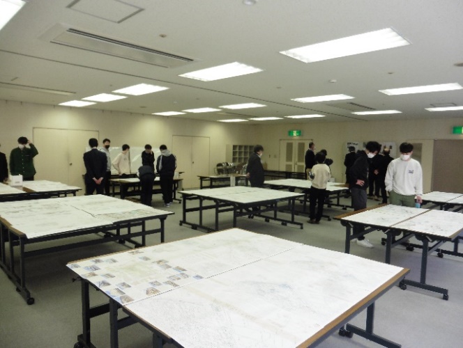 等間隔に設置された机の上に、発表資料が置かれ、資料を見てまわる学生たちを写した写真