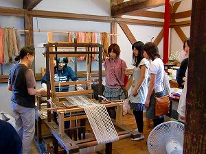教えてもらいながら機織り作業をする大学生と、横で見学する4名の大学生の写真