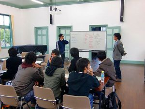 文字が書かれたホワイトボードの前で向き合って話す男性2名と長机に座って話を聞く大学生たちの写真