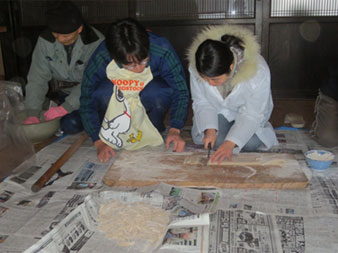 床に敷かれた新聞紙の上で、左側のエプロンをした男性が見守る中、白いコートを着た女性がまな板にのせたうどんの生地を包丁で切っている写真