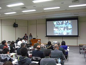 スクリーンに映し出された資料を見ながら発表している2名の女性と、パイプ椅子に座って話を聞く発表会参加者の写真