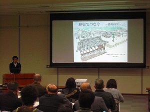 スクリーンに映し出された資料を見ながら発表している男性とパイプ椅子に座って話を聴く発表会参加者達を後方から写した写真