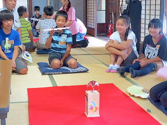和室で紺色の座布団に座った男の子が正座をして扇子を持ち、赤絨毯の上に設置された「枕」と呼ばれる土台に「蝶」と呼ばれる的を立て、蝶へ向けて扇子を投げようとしているのを周りで見ている子ども達の写真