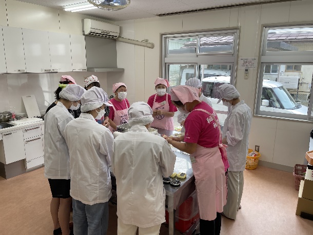 調理室で白衣と白い帽子、マスクをつけた学生たちと、ピンクのエプロンに三角巾、マスクをつけた数名の女性が調理台を囲んで作業をしている様子の写真