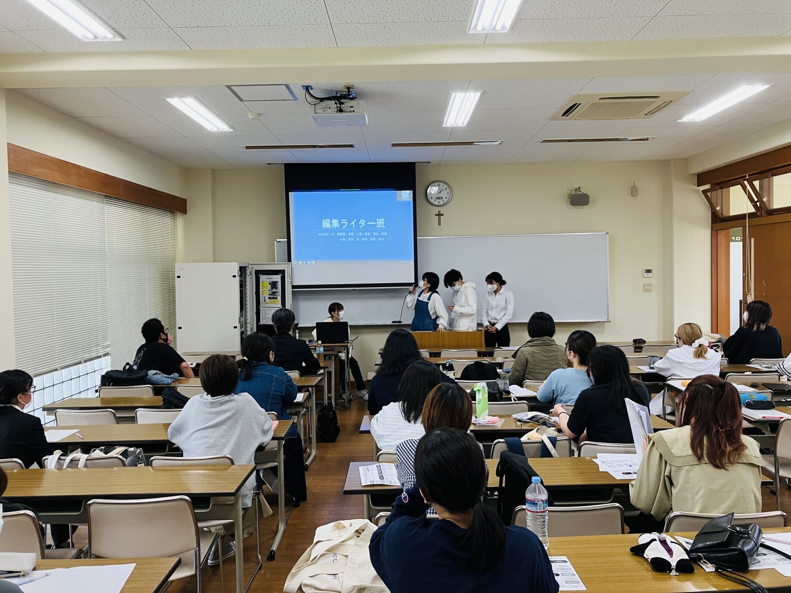 長机が並べられた部屋に多くの参加者が座り、前方のプロジェクターに映し出された映像を見ながら話をしている女性を室内の後方から写した写真