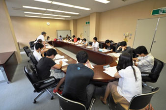 楕円形に設置された茶色い長机に資料を広げ座り、参加者たちが話し合いを行っている様子の写真