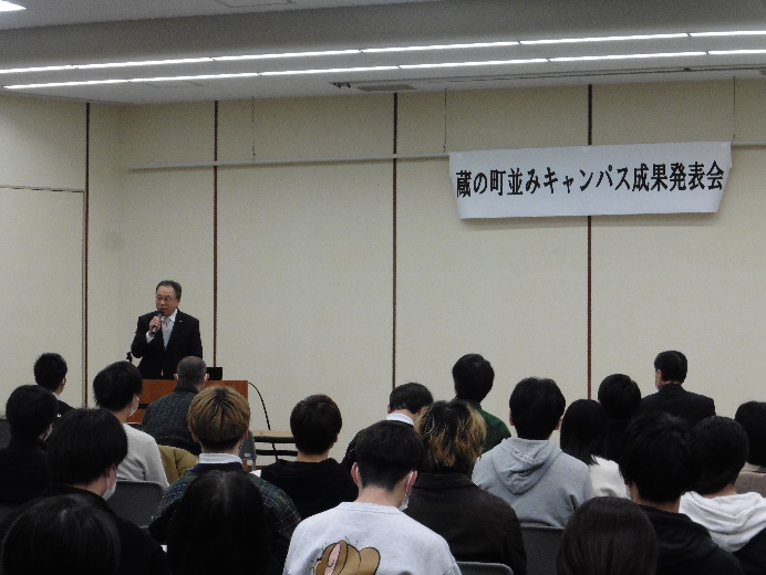 蔵の町並みキャンパス発表会で、前方の演台に立ち、マイクを持って話をしている男性と、多くの参加者を室内の後方から写した写真