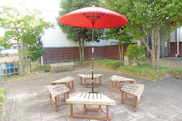 笠鉾会館入り口横の広い場所に設置されたベンチ。台形で、背もたれのない椅子が6個、六角形の形に並んでいる。6つの椅子の中心部には赤い和風のパラソルが立っている。