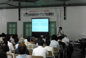 旧小田切家住宅オープニング記念講演会と書かれた横断幕が設置された下で、モニターを使い、その右側でマイクを持った人が参加者の方たちに向かって話をしている様子の写真