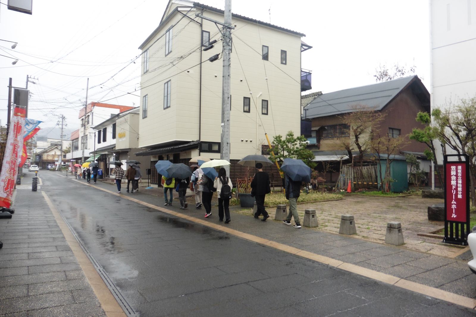 雨の中、歩道を傘をさして歩いている大学生たちの様子を左斜め後ろから写した写真