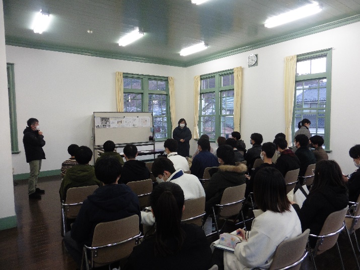椅子に座っている多くの学生たちの正面のホワイトボートに資料が貼られ、左横に立っている男性が資料を見ながら話しをしている様子を学生たちの後方から写した写真