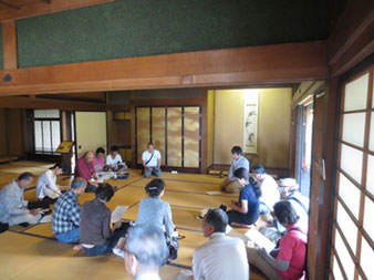 右奥に掛け軸、左に金色の襖がある広い和室で、参加者たちが手に資料を持ち、円になって座り話し合いが行われている様子の写真