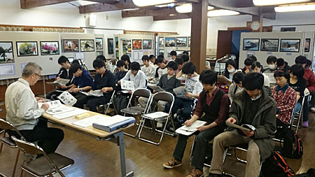 多くの写真が展示されている室内で、作業服の男性が、椅子に座っている大勢の学生達に向かって授業をしている様子の写真