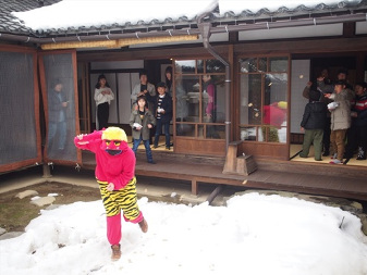 雪が積もっている庭に、虎柄の黄色い短パンを履いた赤鬼の恰好をした人がおり、その鬼に向かって縁側から複数の子どもと大人が豆を投げている節分の写真