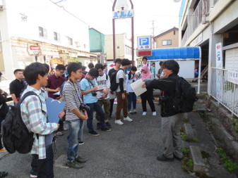 パルムと書かれた看板の近くの駐車場内で、黒い服に黒いリュックを背負った男性が、横3列位に並んだ長野工業高等専門学校 建築設計製図5年生の生徒達に紙を持った手を前に出し話している授業の様子の写真