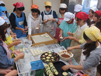 新聞紙が敷かれた卓上に計りやボールなどが置かれ、その周りをエプロンやカラフルな三角巾を身に着けている大学生達が囲み、おやきを作っている写真
