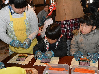 机の上にタッパの中に入った細長くカットされた人参が数本と具材が置かれ、その近くで恵方巻を作っている子ども3人の節分パーティの様子の写真