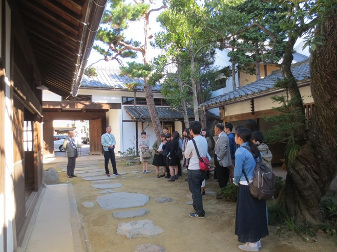 日本家屋に囲まれた飛び石のある庭で、作業服とスーツを着た男性2人の説明を聞く多文化コミュニケーション学科日本語日本文化専攻の人達の研修の様子の写真