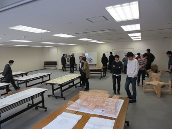 資料が置かれた長机が2列3台ずつ広い室内に置かれており、資料を見ているスーツを着た男性、白シャツにジーンズを着て話をしている2人、奥でホワイトボードを見ている人達などが写っている蔵の町並みキャンパス成果発表会の様子の写真
