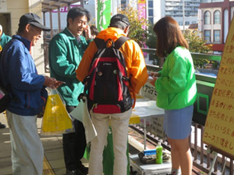 須坂駅にて左から黒い帽子を被り青いジャケットを着た男性、緑のジャンパーを着た男性、オレンジ色の上着を着て赤と黒のリュックを背負った男性と、一番右に蛍光緑の上着と水色のスカートを履いた女性が話している様子の写真