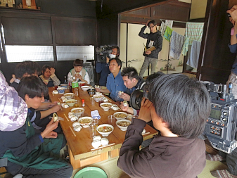 古民家にて美味しそうな食事が並べられた長机を囲み、美味しそうに食べる学生達とその様子を手前でカメラを右肩に担いで撮影している男性が写っている写真