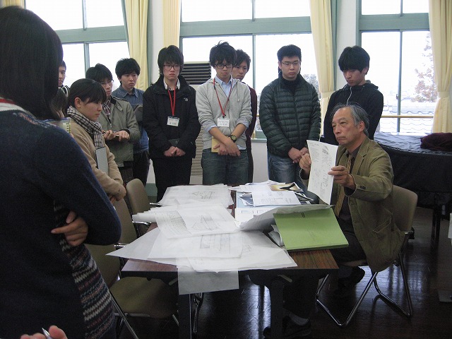 中央にある大量の資料が置かれた机を囲う生徒たちに、草色のジャケットを着た教授が一枚の資料を両手で持ち見せている写真