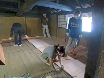 マスクと手袋をして古民家の床に綺麗な板を敷く作業をしている様子の写真
