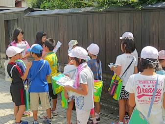 屋外の木製の塀の前で、白い帽子を被りクリップボードを首から下げた子ども達が集まっている写真
