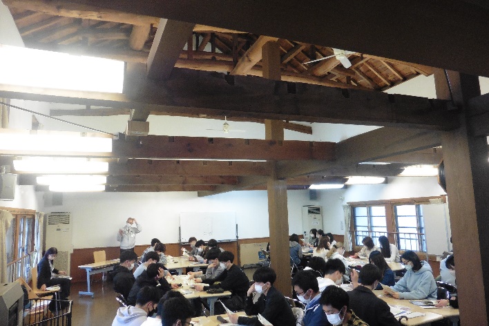 学生たちがグループごとに分かれて席に着き、前方のグレーの服を着た男性が話をしている授業風景の写真