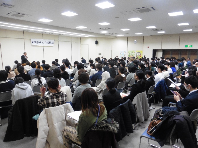 蔵の町並みキャンパス成果発表会で、前方に立ち、話をしている男性と、椅子に座っている多くの参加者を左斜め後ろから写した写真