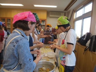 長方形の机を囲う様にエプロンと三角巾を着けた生活科学科健康栄養専攻の女生徒達がおやきの生地を手でこねたり成形している様子の写真