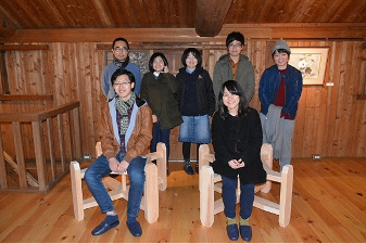 木造の建物内で壁側に笑顔の5人の男女が立ち、その前に製作ベンチの椅子に座ってカメラ目線の2人が写った信州大学工学部建築学科建築心理学の学生の集合写真