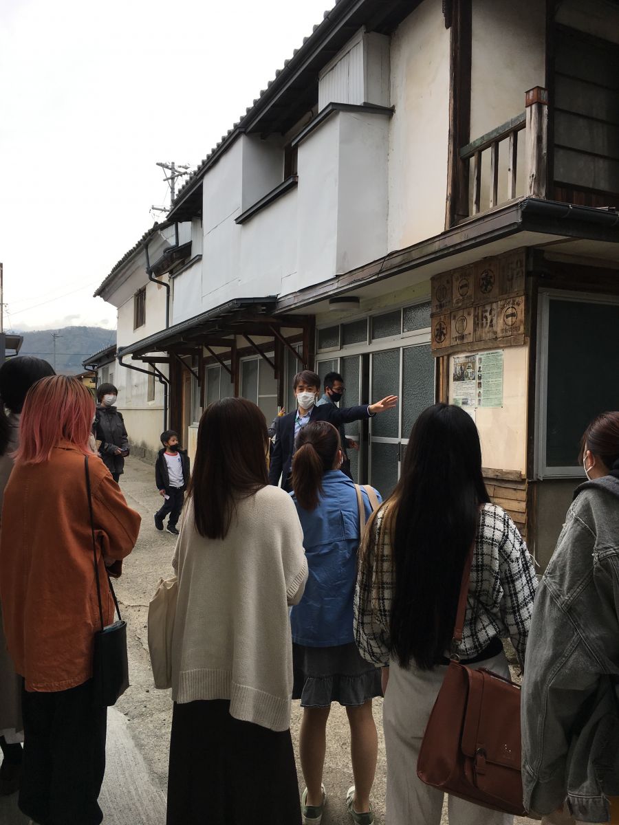 2階建ての建物の1階部分に掲示された紙を指しながら話をしている男性と、真剣に話を聞いている大学生の様子を大学生の後方から写した写真