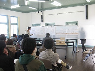 資料が貼られたホワイトボードを指さし棒で差しながら話す男性とパイプ椅子に座って話を聴く大学生の写真