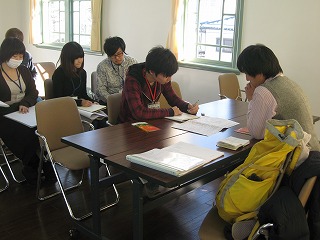 長机を2台並べて向き合って話す男性2名と、その後ろでパイプ椅子に座る4名の大学生の写真