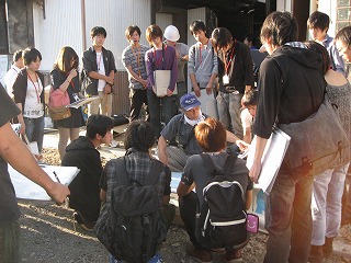 話をする紺色の帽子を被った男性と、男性の周りを囲んで話を聴く大学生の写真