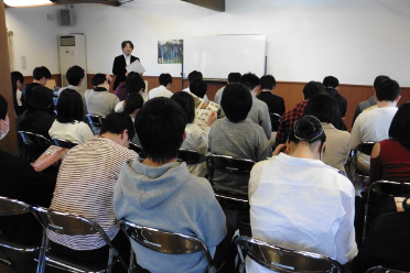 ホワイトボードの左側でスーツを着た男性が資料を持ち、パイプ椅子に座った多くの学生達に話をしている授業の様子の写真
