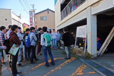 立体駐車場の前で立て看板を指さし話をする黒ジャケットを着た男性と、その周囲にいる学生たちの授業の様子の写真